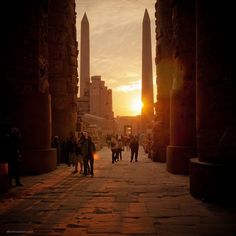 the sun is setting behind two obelisk in an alley way with people walking on it