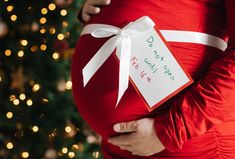 a pregnant woman wearing a red dress and white ribbon wrapped around her belly with a christmas tree in the background