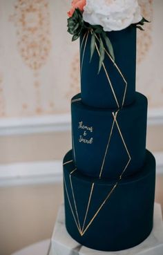 a three tiered blue wedding cake with gold lines and flowers on top is decorated with greenery