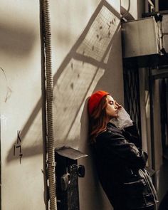 a woman leaning against a wall wearing a red hat and black coat with her eyes closed