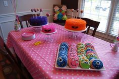 a table topped with cakes and cupcakes covered in frosting on top of a pink table cloth