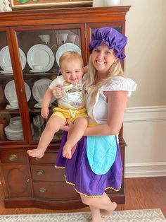 a woman holding a baby in front of a china cabinet with plates on top of it