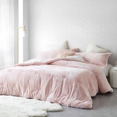 a bed with pink comforter and pillows in a white room next to a window