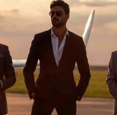 three men in suits standing on an airport tarmac, one is wearing sunglasses and the other has his hands in his pockets