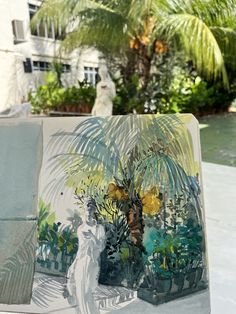 a painting is shown in front of a palm tree and other tropical plants, with a white statue behind it