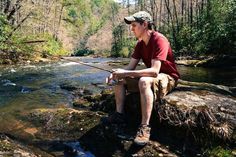 a man sitting on top of a rock next to a river holding a fishing pole