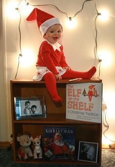 a baby sitting on top of a bookshelf with christmas lights in the background