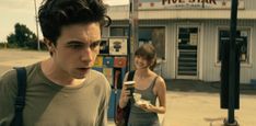 two people standing in front of a store on the side of the road with food