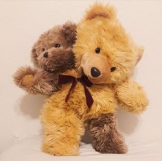 two brown teddy bears sitting next to each other on a white surface with one holding the other