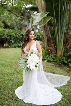 a woman in a wedding dress holding a bouquet and posing for the camera with greenery behind her