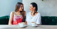 two women sitting at a table with cups of coffee in front of them, talking