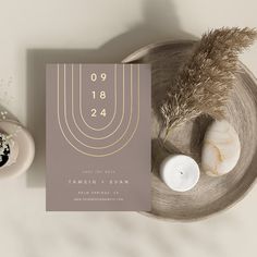 a pink and gold wedding card on a plate next to a candle, some dried flowers and a vase