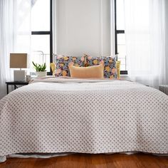 a bed with white sheets and pillows in front of two windows, next to a wooden floor