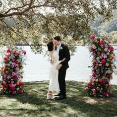 a bride and groom kissing in front of an arch with flowers on the grass near water