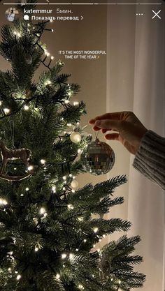 a christmas tree is decorated with white lights and silver bauble ornaments, while someone's hand reaches for the ornament