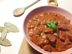 a pink bowl filled with meat and vegetables on top of a wooden cutting board next to a spoon