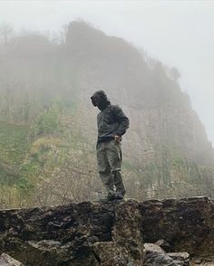 a man standing on top of a large rock near a mountain covered in fog and mist