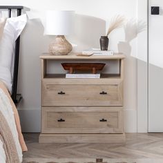 a nightstand with two drawers and a lamp next to it on top of a bed