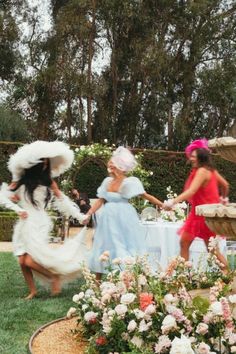 two women in dresses and hats are dancing near a fountain with flowers on the ground