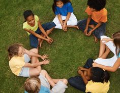 a group of children sitting in a circle on top of grass with their hands together