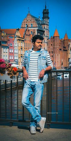 a man leaning on a railing in front of some buildings and water with his hands behind his back