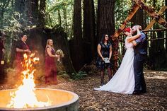 a bride and groom standing in front of a fire pit with their wedding party around it