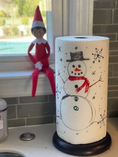 an elf is sitting in the window sill next to a toilet paper roll with a snowman on it