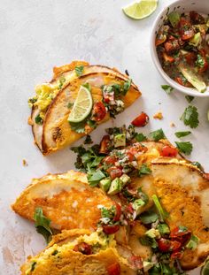 some food is laying out on a white surface next to a bowl of salsa and lime wedges