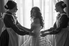 three women in dresses are standing near a window and one is holding the other's hand