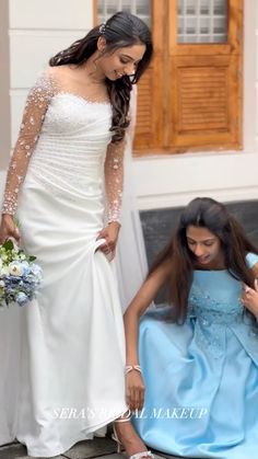 two women in wedding gowns standing next to each other and one woman sitting on the ground