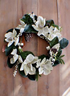 a wreath with white flowers and pine cones on a wooden floor, surrounded by greenery