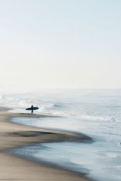 a person with a surfboard standing on the beach near the ocean and looking at the water