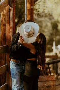 a man and woman hugging each other with a cowboy hat on their head, leaning against a wooden fence