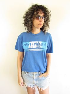 "Details:  Vintage 80s Sun Sportswear T shirt Blue with front decal of a Sailboat and beach/ palm trees single stitch/ crewneck distressed-fading, pilling, decal cracking Brand: Sun Sun Sun Material: Cotton/ Poly Size: M Measurements Approx: taken flat then doubled if needed Shoulders: 18\" Chest: 39\" Sleeve: 7\" Waist: 39\" Length: 25\" Condition: Good Vintage- distressed wear-has fading, holes, pilling, pulls, spots, decal cracking, etc. *Colors of items may differ slightly from photos due to Blue Retro Tops For Beach Season, Relaxed Fit Blue Tops For Surfing, Blue Palm Tree Print Tops For Beach Season, Blue Relaxed Fit Tops For Surfing, Vintage Crew Neck Tops For Beach Season, Casual Blue Palm Tree Print Top, Beachy Crew Neck Top With Palm Tree Print, Blue Surfing Tops With Screen Print, Blue Cotton Top With Palm Tree Print