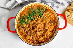 a red pot filled with beans and meat on top of a counter next to bread