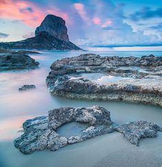 an ocean scene with rocks in the foreground and a mountain in the background at sunset