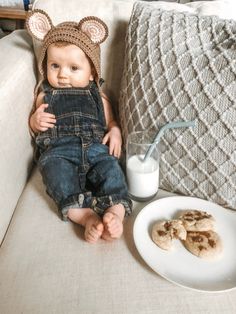 a baby in overalls sitting on a couch next to a plate of cookies and milk