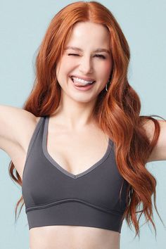 a woman with long red hair wearing a gray sports bra top and smiling at the camera