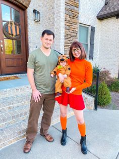 a man and woman standing in front of a house holding a baby wearing an orange outfit