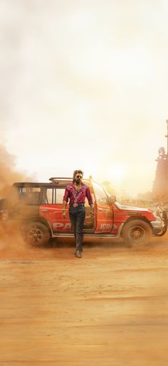 a man standing next to a red truck in the middle of a dirt road near a tall building