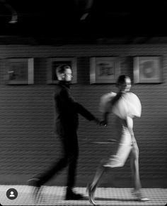 black and white photograph of two people walking in front of a wall with pictures on it