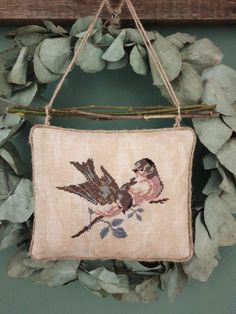 a cross stitch bird hanging from a wooden frame on a wall next to some green leaves