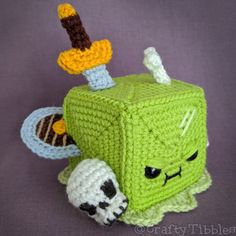 a crocheted green cube with two faces and a skull on the side, sitting in front of a purple background