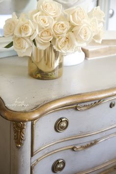 white roses in a gold vase sitting on top of an antique dresser with mirror behind it