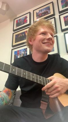 a man sitting in front of a wall with pictures on it playing an acoustic guitar