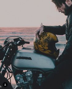 a man sitting on top of a motorcycle next to the ocean and holding a helmet