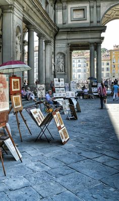 an open air art market with people walking around and looking at paintings on easels