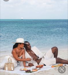 a man and woman sitting on the beach next to each other with food in front of them