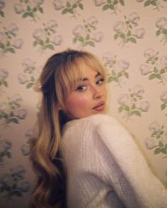 a woman with long blonde hair standing in front of a floral wallpaper