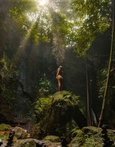 a naked woman standing on top of a rock in the forest with sunlight streaming through the trees